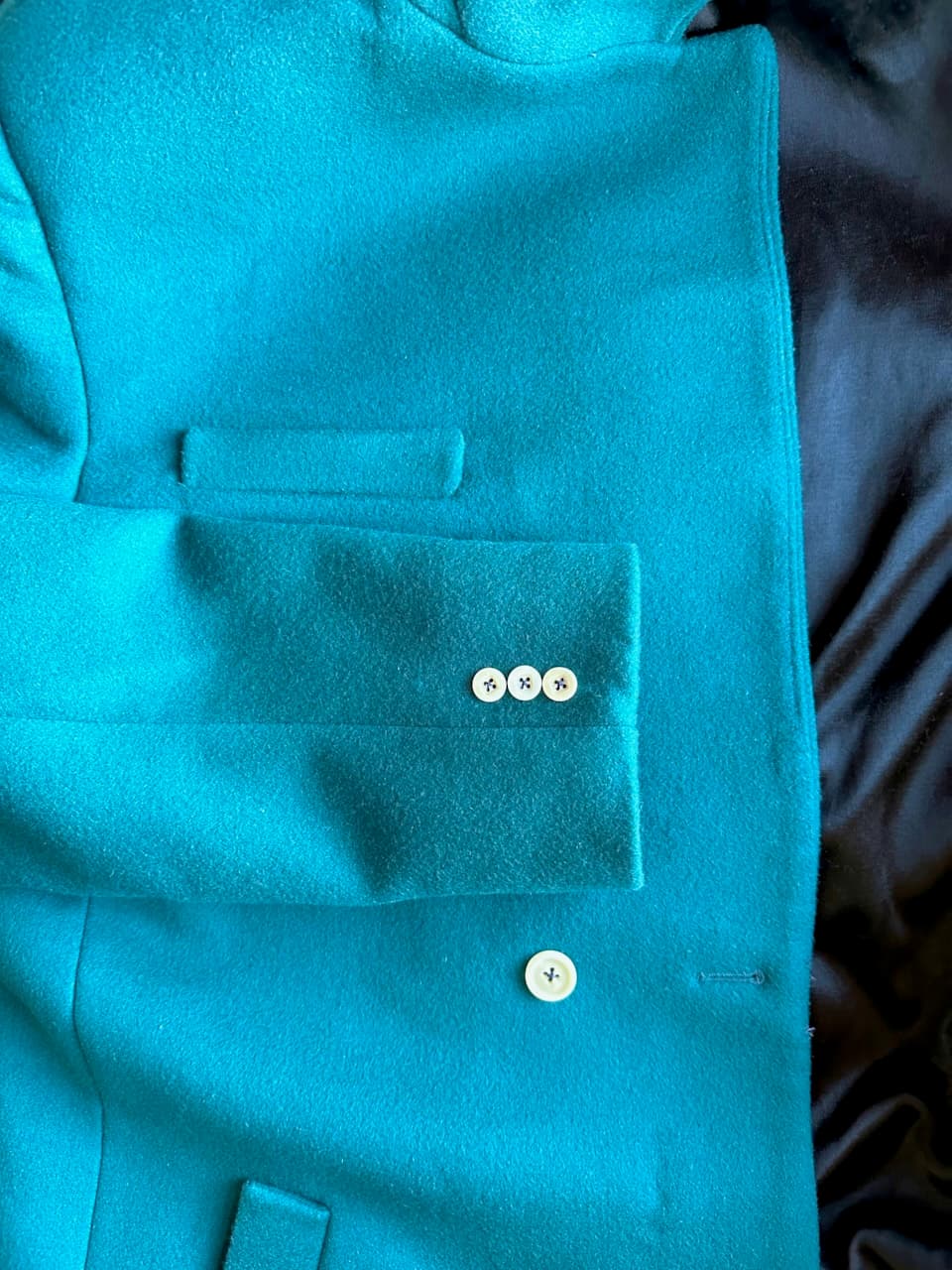 Zoomed in photo of a flat lay of the coat, showing the front button, the matching sleeve buttons, the small horizontal chest welt pocket and the tip of the slanted front welt pocket